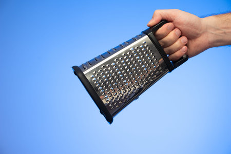 Stainless metal kitchen grater with plastic handle held in hand by Caucasian male hand studio shot...