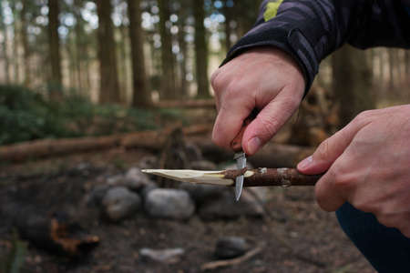 Caucasian male hand in winter jacket sharpening a wooden stick skewer with a nondescript pocket knife close up shot unrecognizable.の写真素材