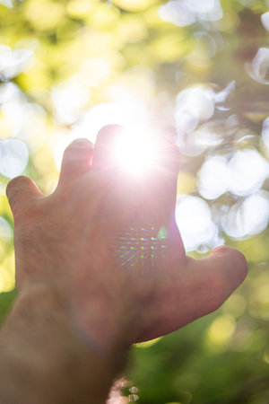 Caucasian male hand reaching upwards open hand close up shot green and yellow nature shallow depth of field background. Reaching out hand gesture. Sun rays flaring through the fingers.の写真素材