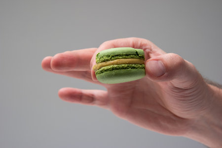 Colorful sweet macaroon cookie held in hand by Caucasian male hand. Close up studio shot, isolated on gray background.の写真素材