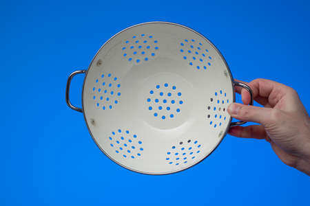 White metal round empty kitchen colander held by male hand. Close up studio shot, isolated on blue background.の写真素材