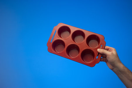 Empty red ceramic muffin pie tray  with cutouts held by Caucasian male hand. Close up studio shot, isolated on blue background.の写真素材