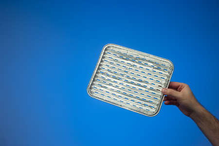 Empty single use metal aluminum foil cooking tray held by Caucasian male hand. Close up studio shot, isolated on blue background.の写真素材