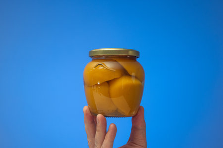 Preserved halved peaches in syrup in a glass jar held by Caucasian male hand. Close up studio shot, isolated on blue background.の写真素材