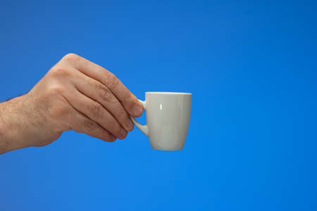 Small white ceramic coffee cup  held by Caucasian male hand. Close up studio shot, isolated on blue backgroundの写真素材