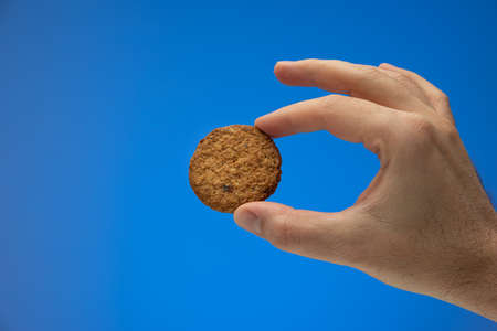 Whole grain brown biscuit or cookie held by Caucasian male hand. Close up studio shot, isolated on blue background.の写真素材