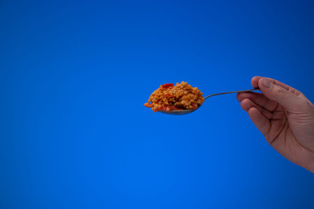 Cooked bulgur with diced tomatoes on a silver spoon held by Caucasian male hand. Close up studio...
