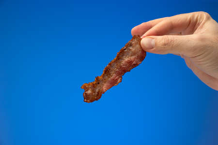 Crispy fried bacon strip or slice held between finger by Caucasian male hand. Close up studio shot, isolated on blue background.の写真素材