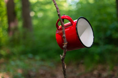 Red metal camping or picnic adventure mug or cup. Day time green forest setting, no people.の素材 [FY310186613239]