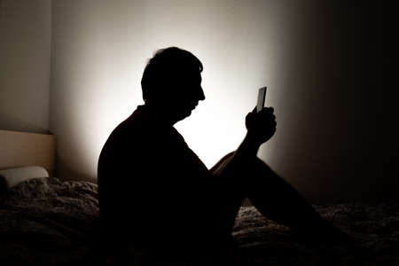Male silhouette depicting loneliness or depression sitting in his apartment bed, looking at social media on his phone.の写真素材
