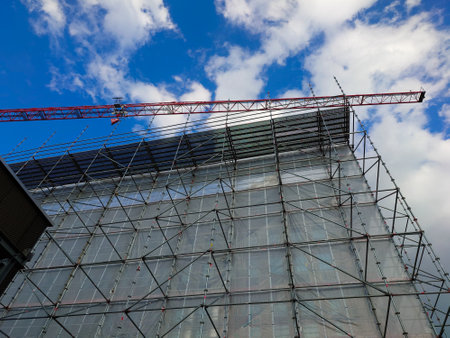 Crane and scaffolding around tall industrial building on a construction site. Low angle view, blue sky, no people.の写真素材