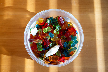 Variety of colored jelly gummy candy in plastic round container. Set on a wooden table, illuminated...