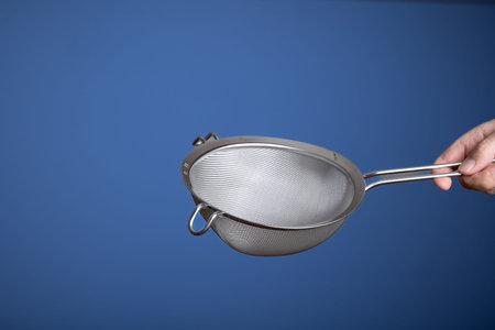 Stainless steel sieve. Held by hand by Caucasian male hand. Close up studio shot, isolated on blue.