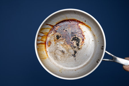 Dirty, oil and grease burnt bits and stains on a metal frying pan. Held in hand by male hand. Close up studio shot, isolated on blue.の写真素材