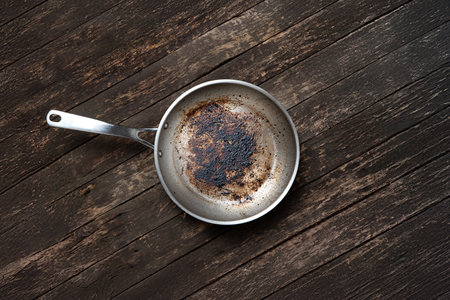 Dirty aluminum or aluminiun fry pan with handle. Burned oily food remains and charred residue inside pan. Top view, wooden background, no peopleの写真素材