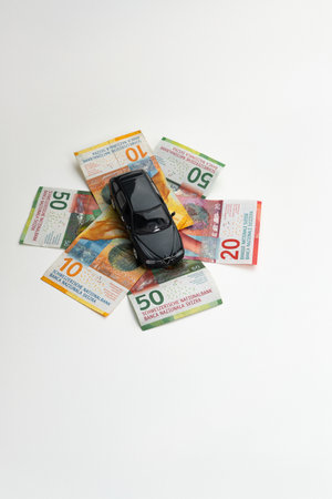 Small metal toy model car on a spread stacks of 10, 20 and 50 banknotes. Top view, close up, isolated on white, no people.の写真素材