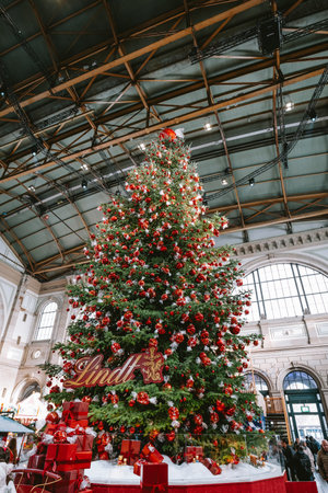 3-12-2024 Zurich, Switzerland. Large Christmas tree inside Zurich main station decorated with red Lindt chocolate balls. Wide-angle, looking up shot, people in the background.のeditorial素材