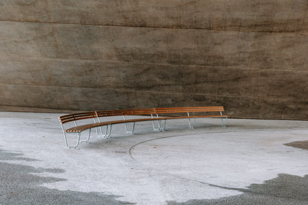Curved, long, empty wooden bench in an urban setting. Concrete floor and walls behind bench, no people.の写真素材