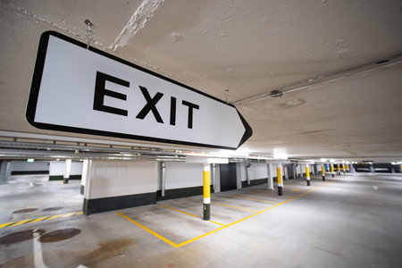 White, large, pointed or directional exit sign inside empty public underground parking lot. Wide...