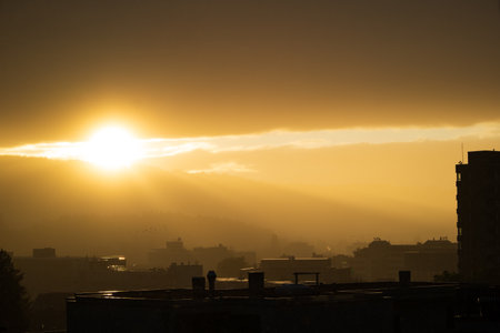 Dramatic sunset silhouette scene. Setting sun piercing behind clouds, city architecture in the foreground, no people.の写真素材