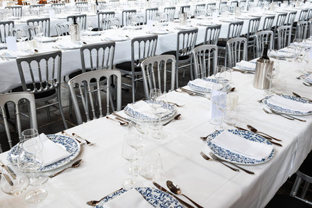 Formal dinning setup of chairs, long tables with tablecloths, plates, cutlery and glasses. Wide...