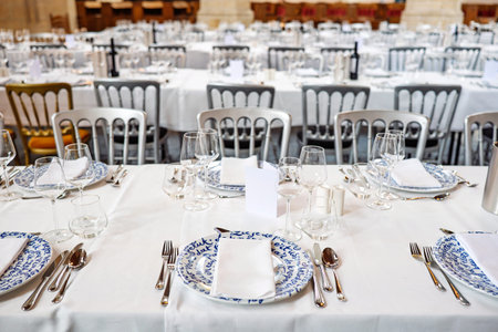Formal dinning setup of chairs, long tables with tablecloths, plates, cutlery and glasses. Wide...