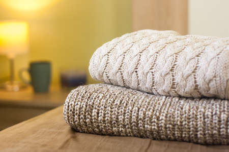 Stack of knitted sweaters on a bed. Small lamp and cup on the wooden table in the background. Warm cozy concept.の写真素材