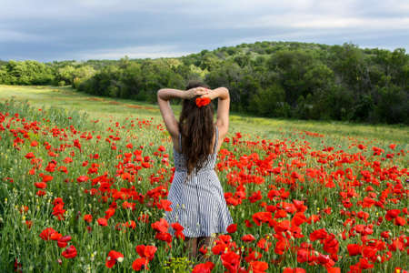 Young woman standing in the middle of vibrant field of red poppies. Cloudy dramatic sky.の写真素材