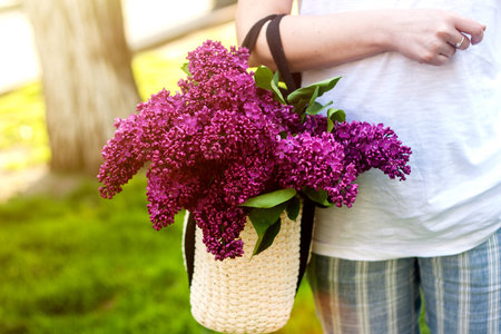 Stylish woman holding straw bag with a vivid bunch of lilac flowers. Bright green grass background.の写真素材