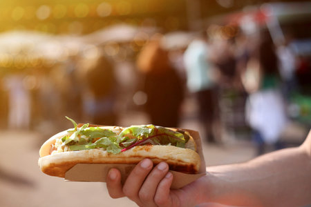 Close up of a hot dog in hand against blurred summer street food festival. Sun flare, defocused people.の写真素材