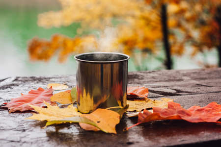 Hot drink in the steel cup on the wooden table. Autumn orange leaves. Yellow trees and lake in the background.の写真素材