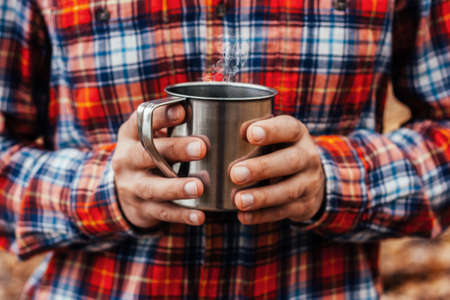 Steel cup with a hot drink in man's hands in the autumn park. Fall background.の写真素材