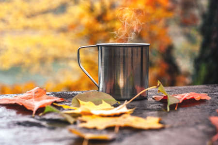 Hot drink in the steel cup on the wooden table. Autumn orange leaves. Yellow trees and lake in the background.の写真素材