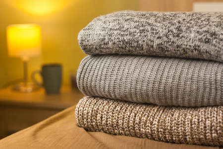 A pile of knitted winter or autumn sweaters on the wood bed. Lamp and cup in background. Warm comfortable clothes.の写真素材