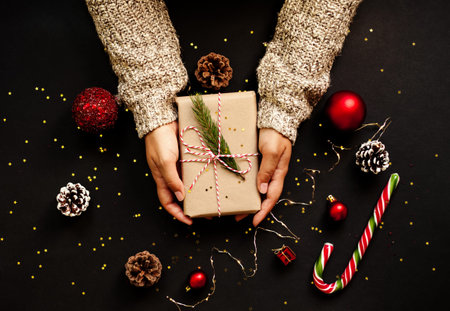 Christmas craft gift boxes with red ribbons, pine cones and red and golden decorations against black background. Gift in female hands.の写真素材