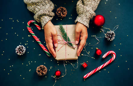 Christmas craft gift box with red ribbons, pine cones and red and golden decorations against dark blue background. Gift in female hands.の写真素材