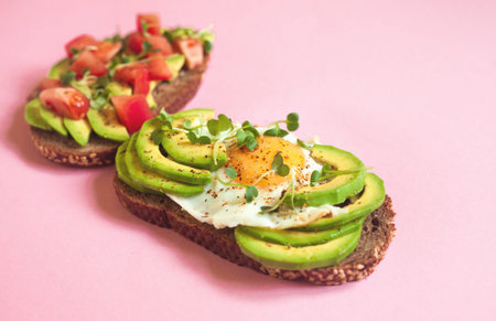 Two toasts of dark bread with avocado slices, chopped tomatoes, fried egg and microgreen. Breakfast concept. Top view, pink background. Healthy food.の写真素材