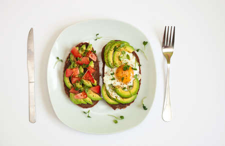 Two toasts of dark bread with avocado slices, chopped tomatoes, fried egg and microgreen. Breakfast concept. Top view, white background. Healthy food.の写真素材