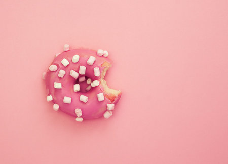 Pink frosted eaten donut with white marshmallows on pink background. Top view, flat lay, copy space.