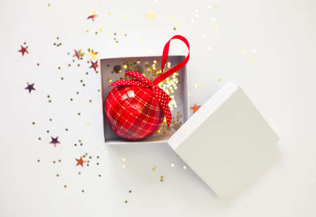 Red Christmas ball in white gift box with cover and golden star confetti on white background. Festive flat lay.の写真素材