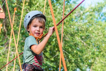 Rope climbing center.Walking in protective equipment. Baby boy playing in adventure Park passes obstacles.Extreme children's leisure and entertainment. Sports are for the nimble and the bold. Portraitの写真素材