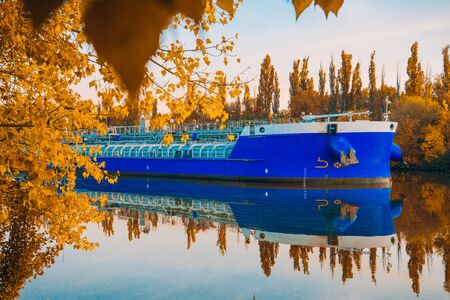 Beautiful autumn evening landscape. Colorful forest and ship quietly floating on the river. Blue barge on a background of Sunny leaves. Indian summer. Yellow trees in the rays of the departing sunの写真素材