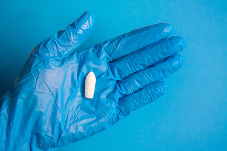 White suppository for anal or vaginal use in hand of a doctor wearing gloves on a blue background....