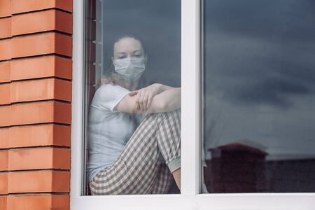 Home quarantine. Caucasian woman sitting at window in a medical mask, looking out, wants to go out. protection against coronavirus infection, pandemics, disease outbreaks and epidemics. Copy space.の写真素材