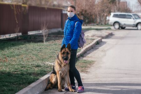 A girl in a protective medical mask walks a dog on the street. Leisure with a pet during quarantine. Walk with a German shepherd in the fresh air. Self-isolation and protection mode.の写真素材