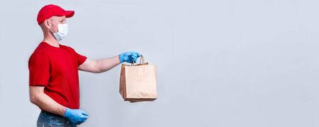 The concept of safe delivery. Banner. A courier in a red uniform and protective medical mask and gloves holds a paper bag, contactless delivery of orders in quarantine during the coronavirus pandemic.の写真素材