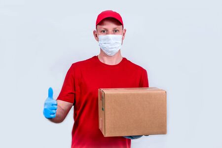 Safe delivery. Courier in red uniform and protective medical mask and gloves holds a cardboard box, contactless delivery of orders in quarantine during coronavirus pandemic. Donation of volunteers.の写真素材