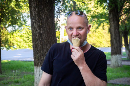 A Caucasian man in a protective mask walks down the street and eats ice cream. Concept of summer and...