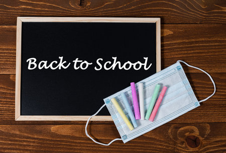 A blackboard with text Back to School, set of colored crayons and a protective mask on a wooden...