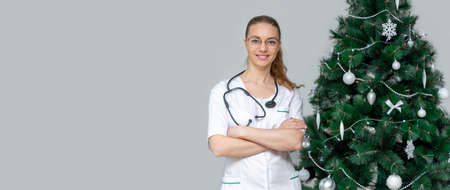 Banner. female doctor in a white uniform smiles next to a Christmas tree. Congratulations to medical professionals on new year and Christmas. New years holidays in the hospital. Copy space for textの写真素材